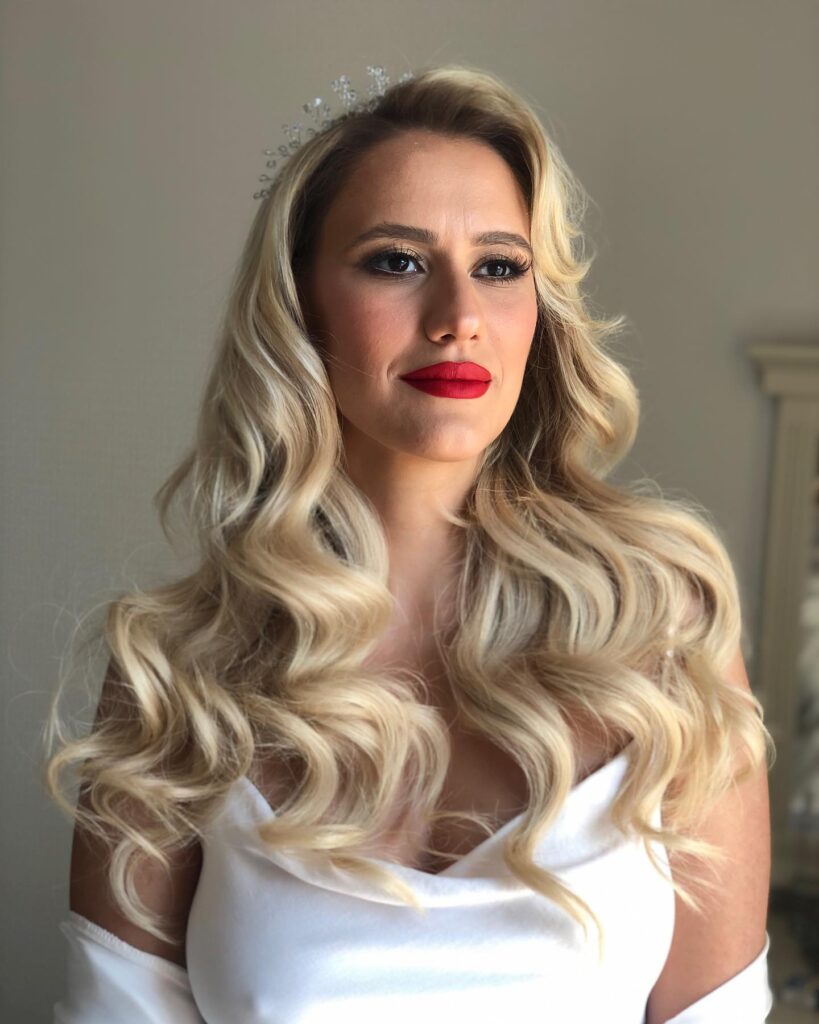 A woman with long, champagne blonde hair styled in voluminous retro waves with a side part and a crystal tiara.