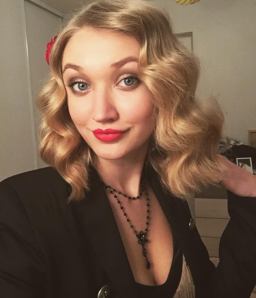 A woman with shoulder-length golden blonde hair styled in soft vintage finger waves with a red flower hair accessory.