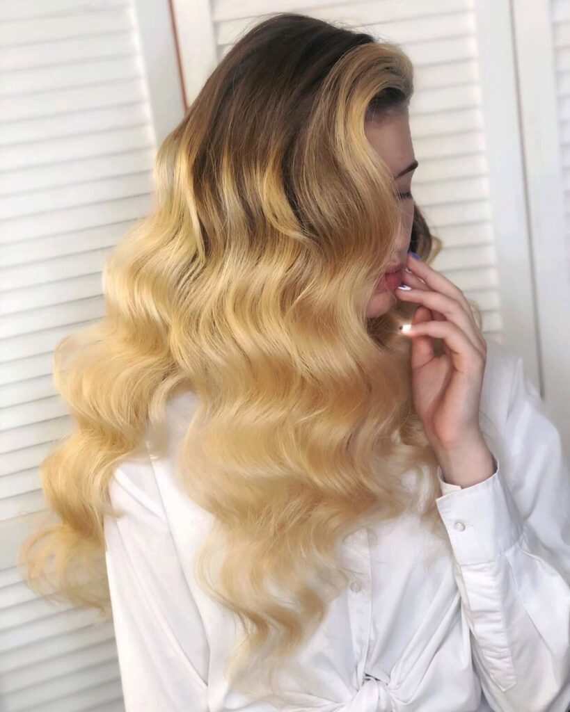 A woman with long ombre hair transitioning from brown to golden blonde, styled in voluminous and uniform vintage waves.