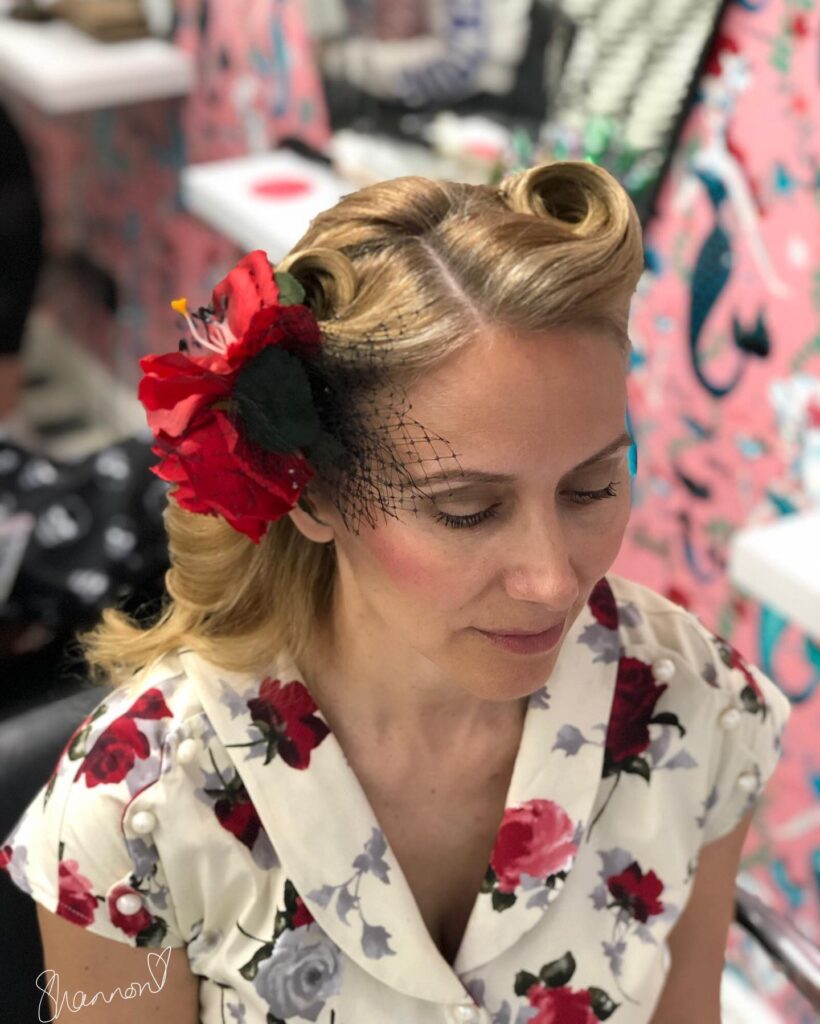 A woman with blonde hair styled in two vintage victory rolls decorated with a large red flower and a black birdcage veil.
