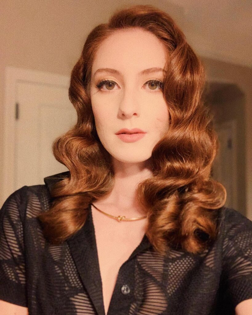 A woman with medium-length auburn red hair styled in glossy and uniform vintage Hollywood waves.