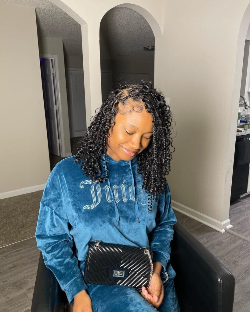 A woman smiling while displaying short black cornrows that flow into voluminous shoulder length curly extensions with detailed baby hairs.
