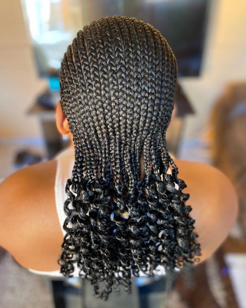 A back view of a person wearing sleek jet black cornrows that end in thick glossy spiral curls at the mid back.