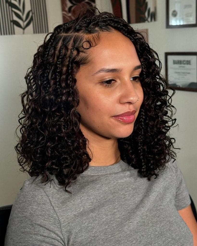 A woman with shoulder length dark curls featuring thin cornrows at the top of her head and a single accent curl on her forehead.