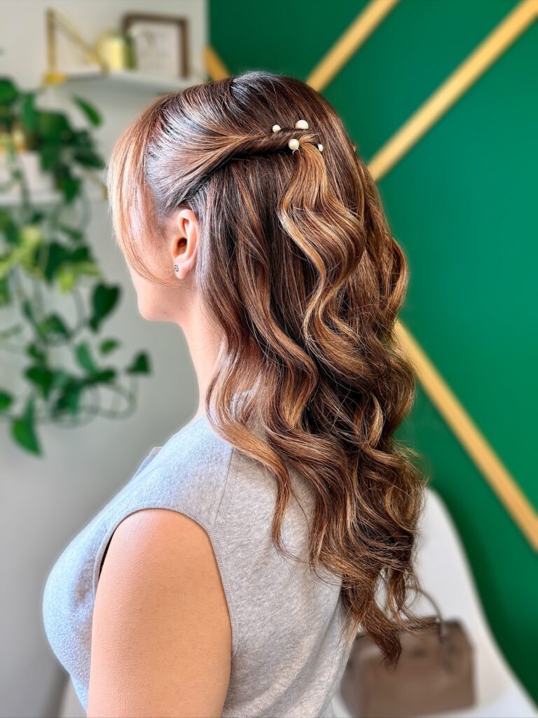 A woman with wavy brown hair with highlights styled in a half up look with side twists and pearl hair pins.