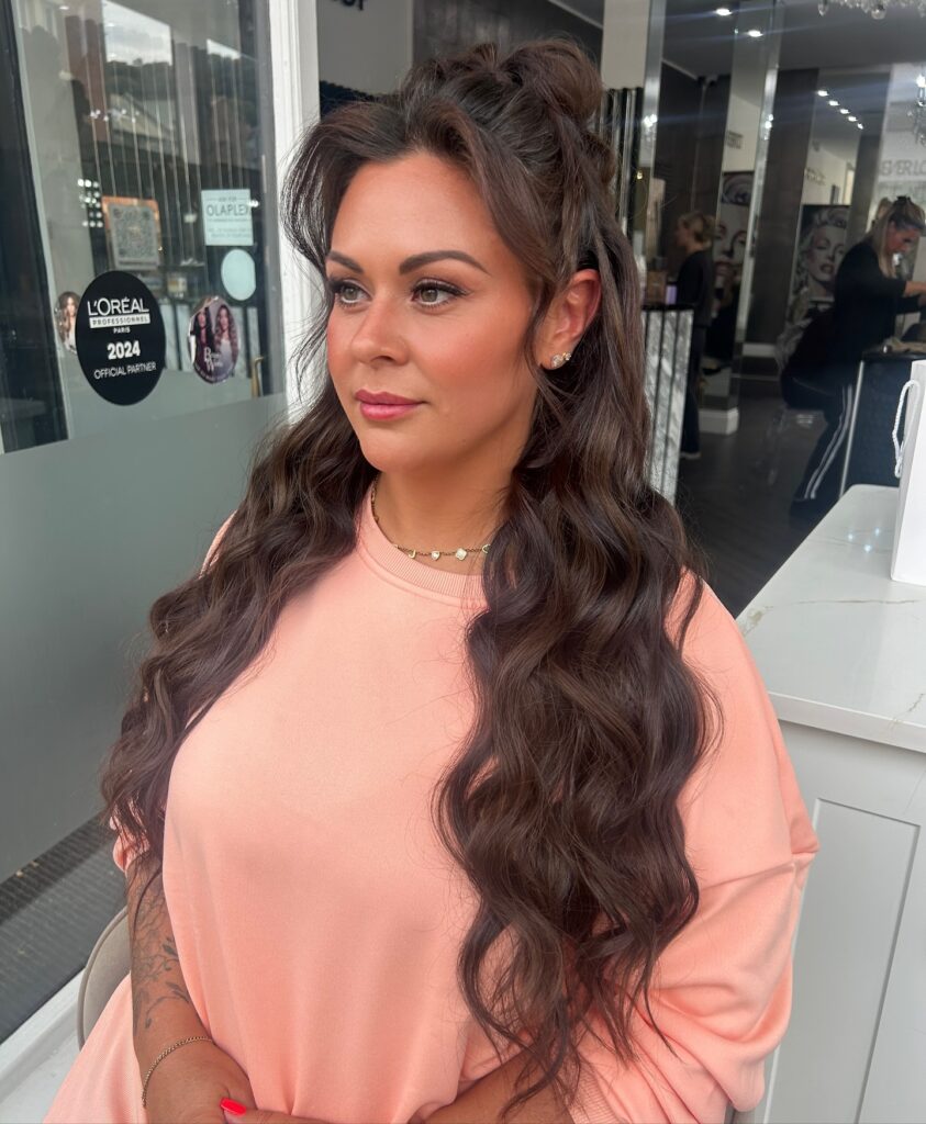 A woman with long wavy dark brown hair styled in a high messy bun half up look with face framing pieces.