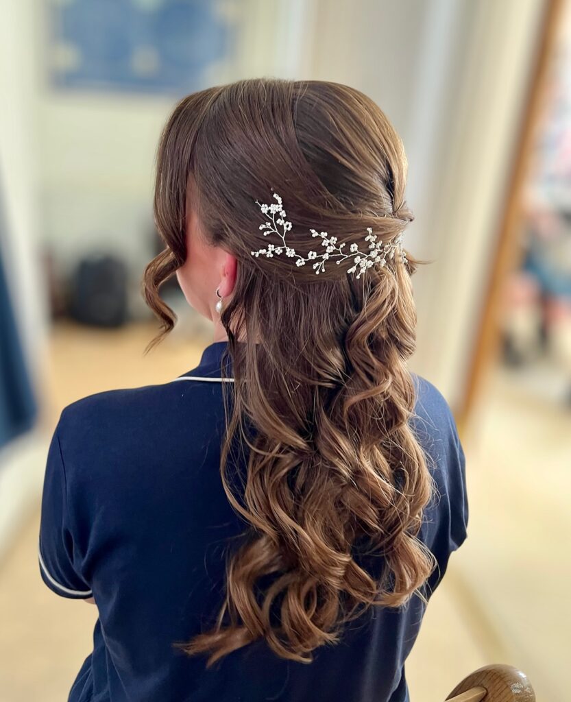 A woman with long wavy brown hair styled in a half up look adorned with a delicate white floral vine hairpiece.
