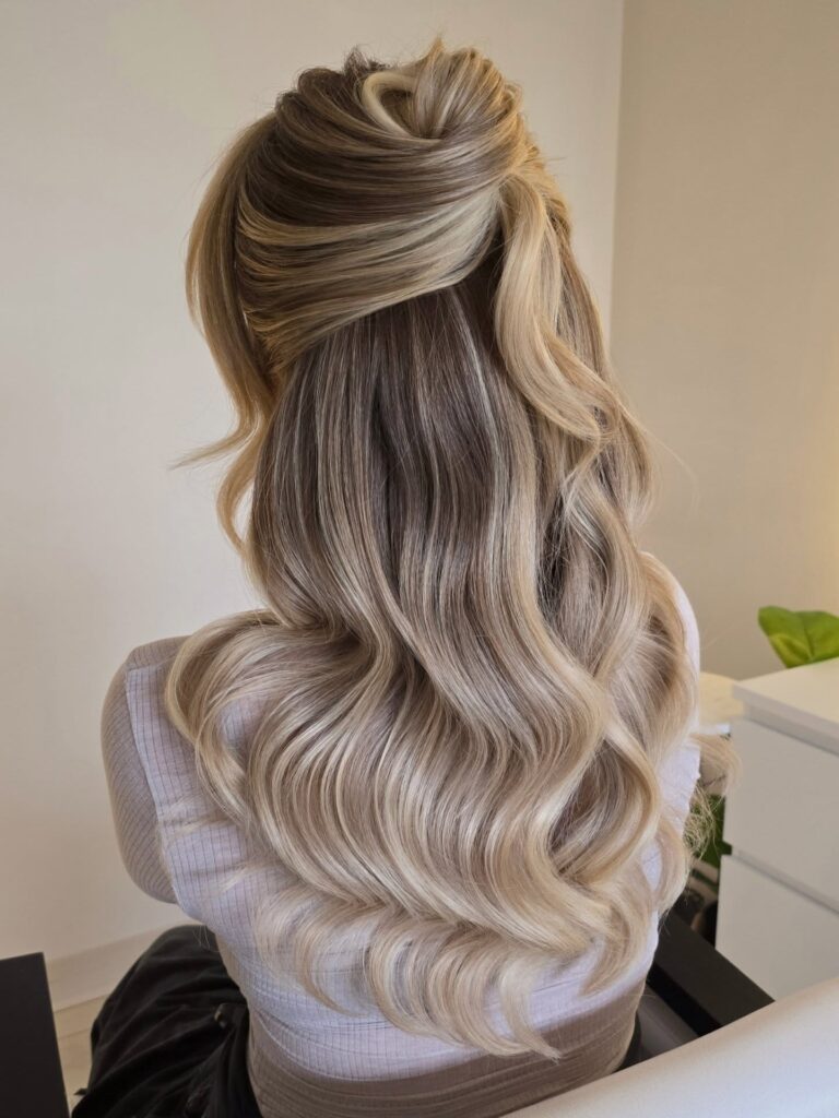A woman with long wavy dark-rooted champagne blonde hair styled in a textured half up hairstyle.