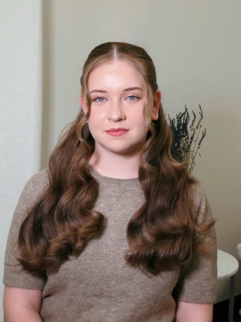 A woman with long wavy brown hair styled in a middle part half up look with two curled strands framing her face.