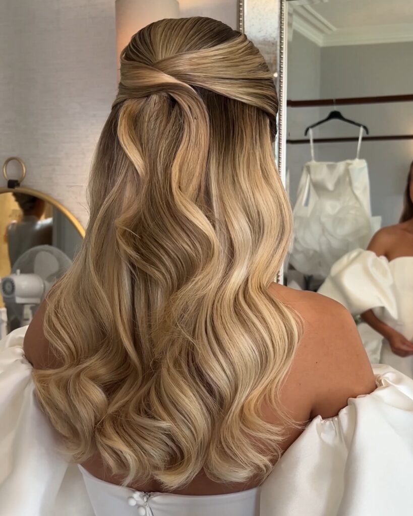 A woman with long honey blonde hair styled in an overlapping half up design with voluminous waves at the ends.