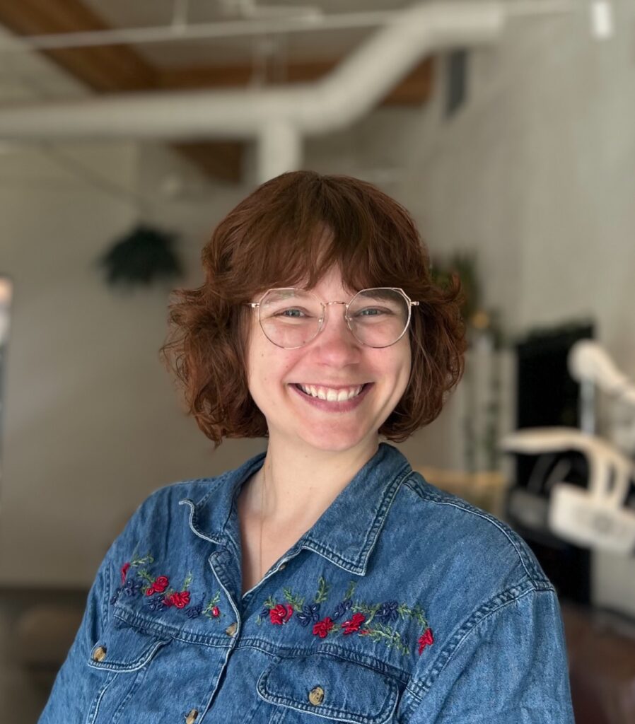 A woman with a curly long bob featuring soft bangs and warm auburn brown tones.