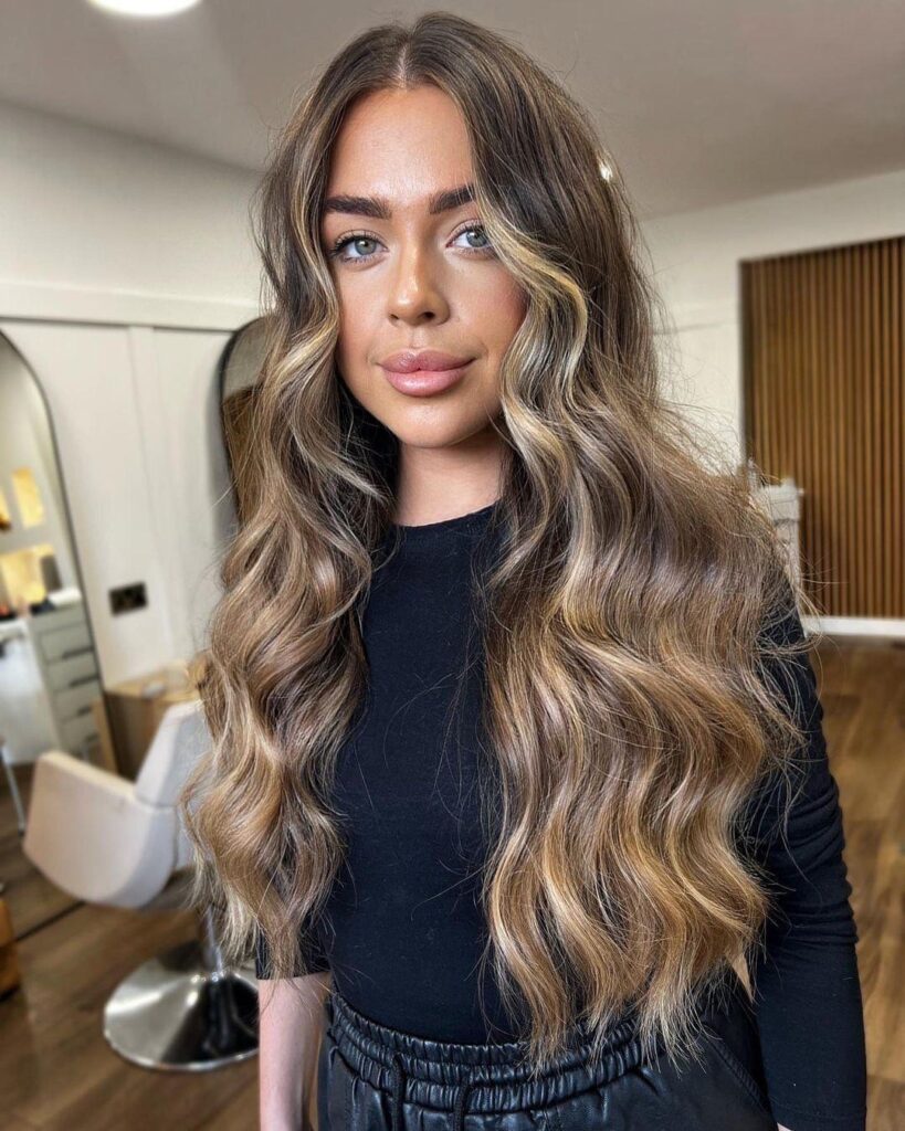A woman with long wavy bronde hair featuring bright face framing highlights and effortless volume.