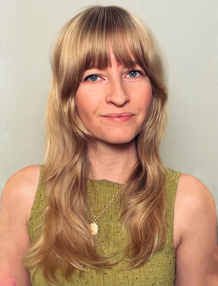 Long blonde layered hair with full feathered bangs and softly curved ends.