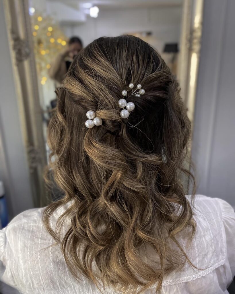 A rear view of a woman with highlighted brown hair styled in half-up twists and loose waves, and adorned with pearl accessories.