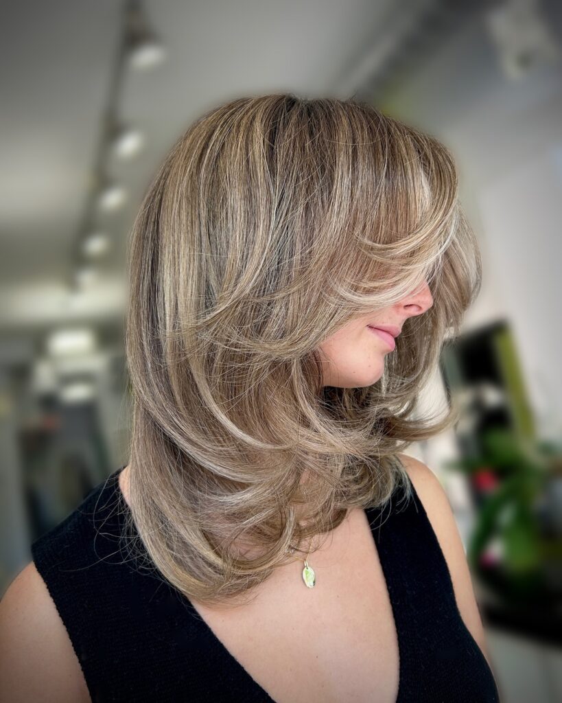 A woman with medium-length ash-blonde hair featuring voluminous butterfly layers and face-framing bangs.