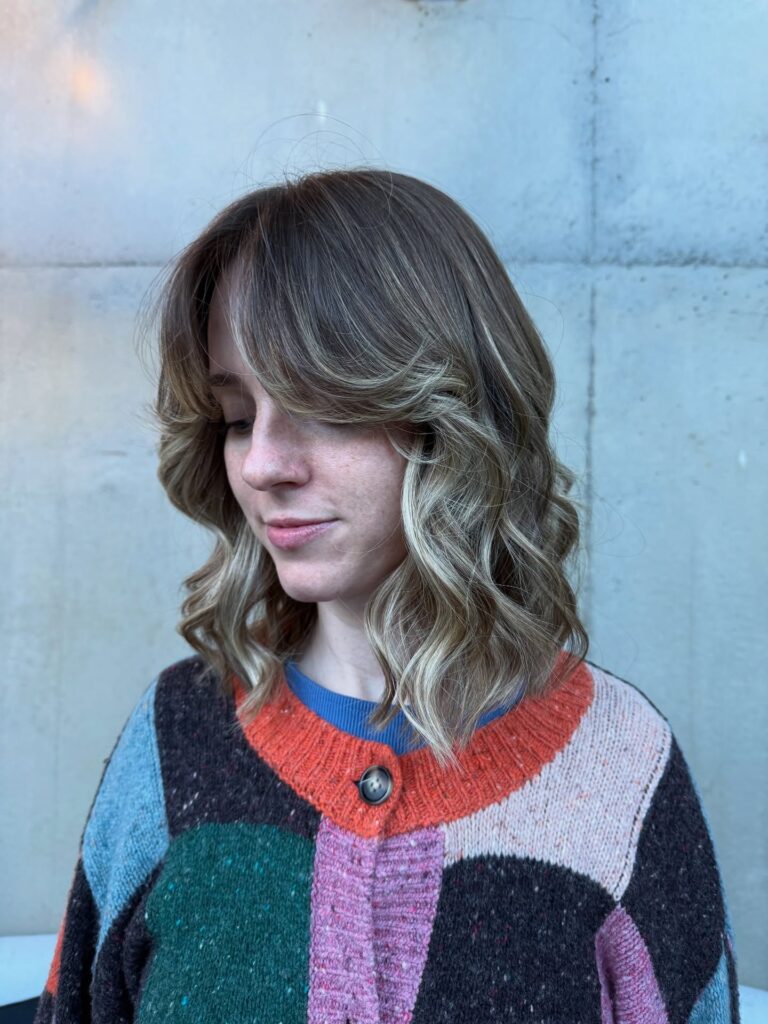 A woman with medium-length blonde hair featuring soft, curtain bangs and textured loose waves.