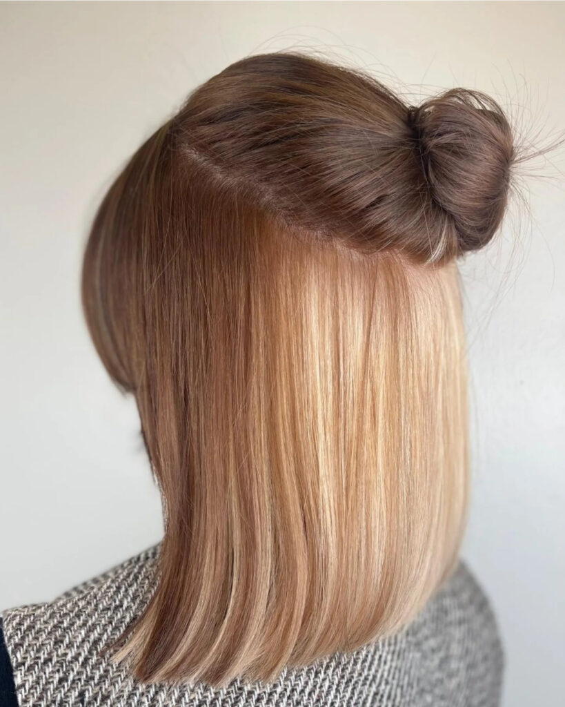 A rear view of a woman with medium-length brown hair styled in a half-up bun, revealing a bright blonde peekaboo layer underneath.