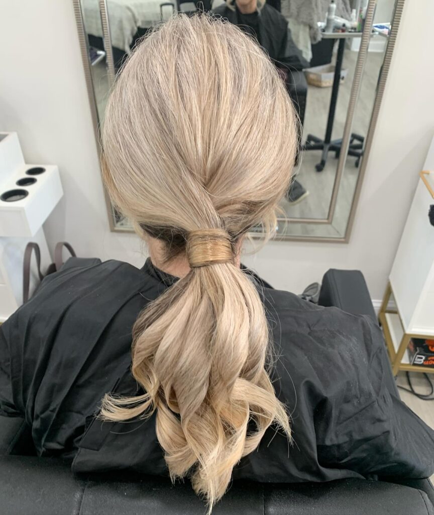 A woman viewed from behind with ash-blonde hair styled in a low ponytail featuring a thick hair wrap around the base and bouncy curled ends.