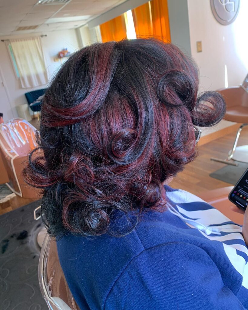 A woman with neck-length black hair featuring bright cherry red highlights and large, bouncy, sculpted curls.