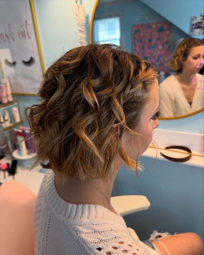 A woman with a chin-length caramel blonde bob featuring loose waves and a small braid along the front hairline.