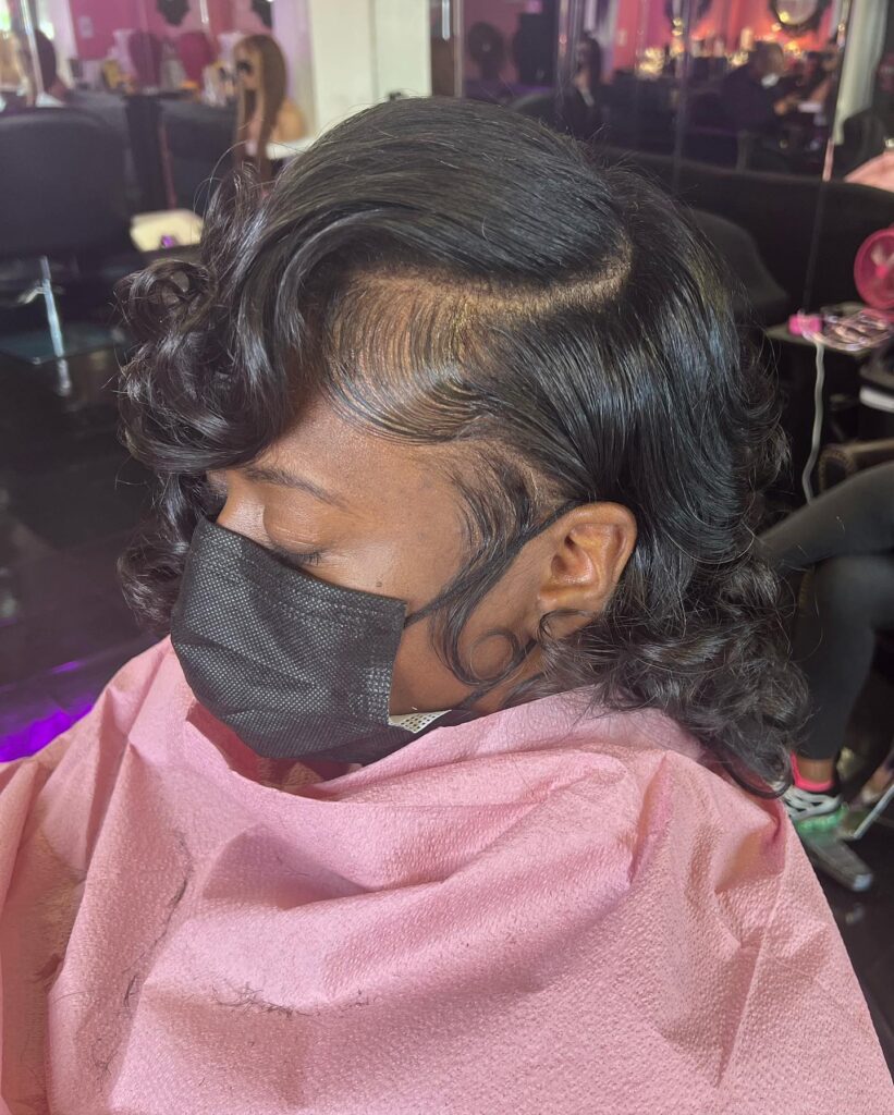 A woman with a neck-length jet-black hairstyle featuring glossy pin curls and decorative laid edges along the hairline.