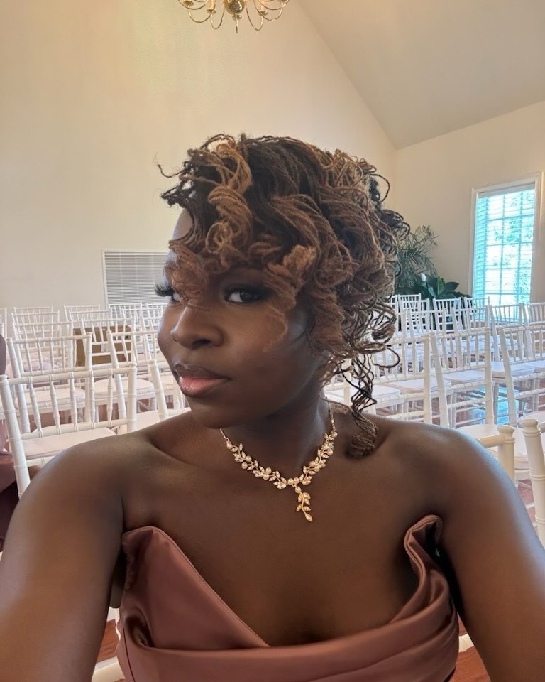 A woman wearing a mauve dress and a gold necklace with her honey-toned micro locs styled in a voluminous, curly updo.