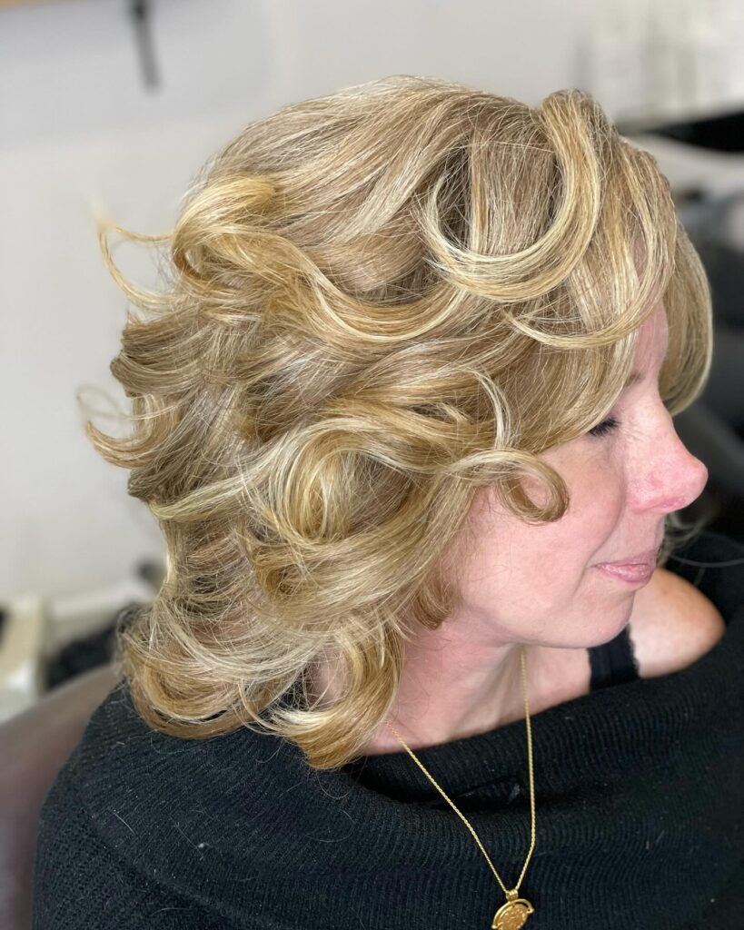 A woman with neck-length honey blonde hair styled in voluminous feathered curls that flick outward.