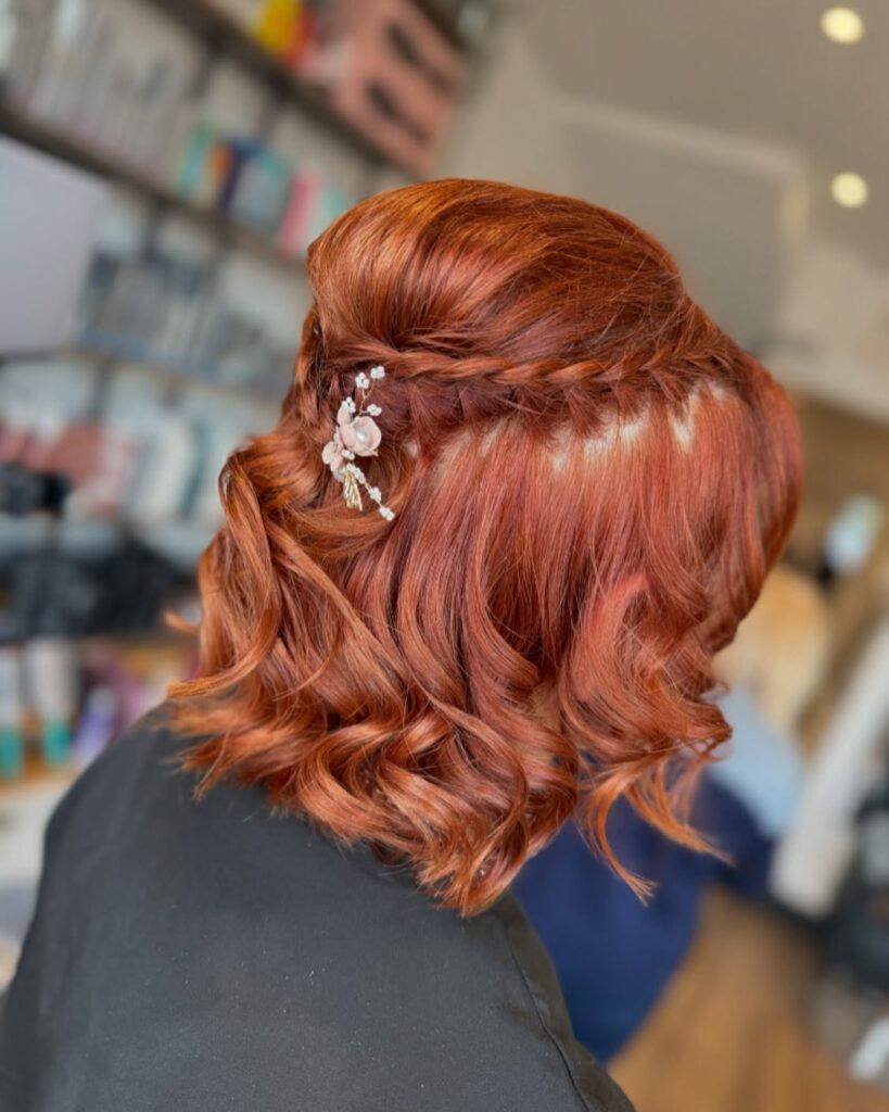 A person with shoulder-length copper red hair styled in barrel curls with a horizontal crown braid and a small pink floral hair clip.