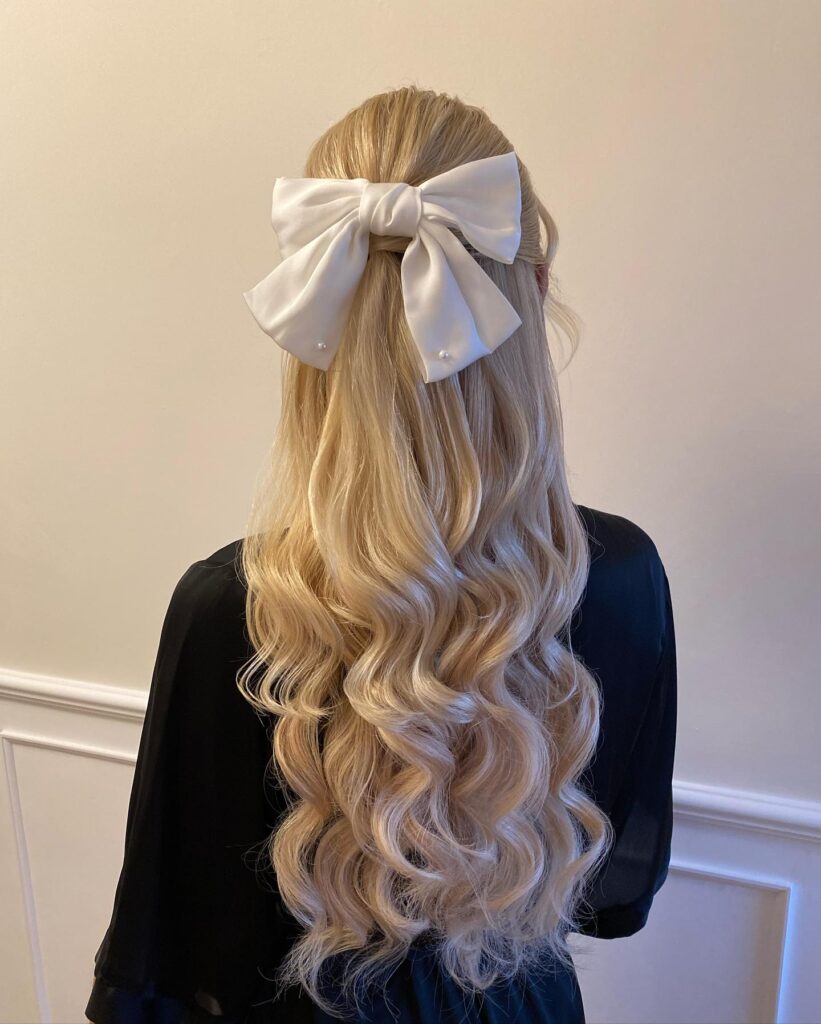 A woman with long wavy blonde hair in a half updo styled with a large white satin bow embellished with small pearls.