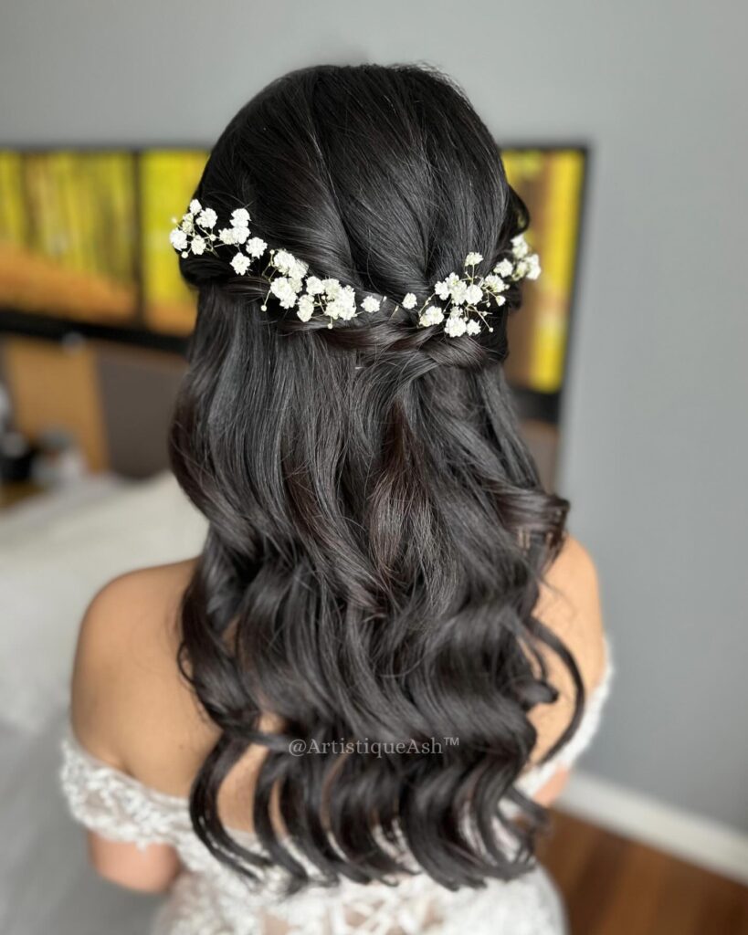 A woman with long, wavy dark brown hair in a half updo decorated with small white flowers along the back.