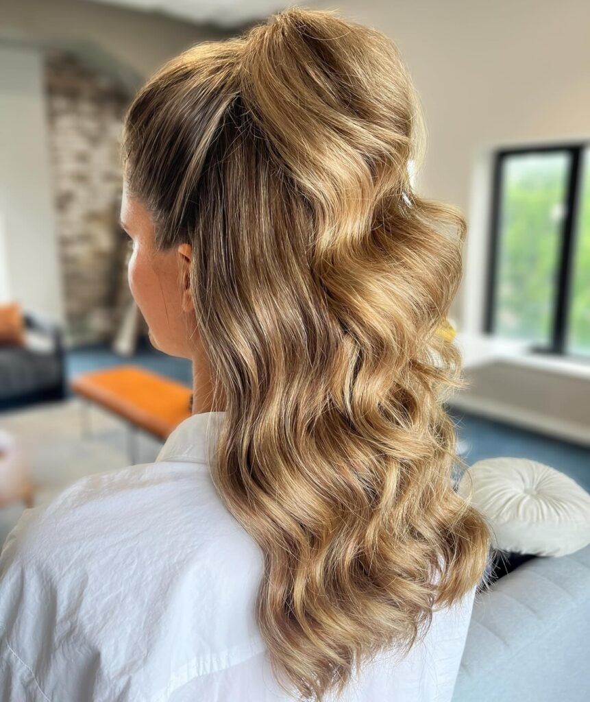 A woman with long wavy ash-blonde hair in a high half-ponytail style with lots of volume.