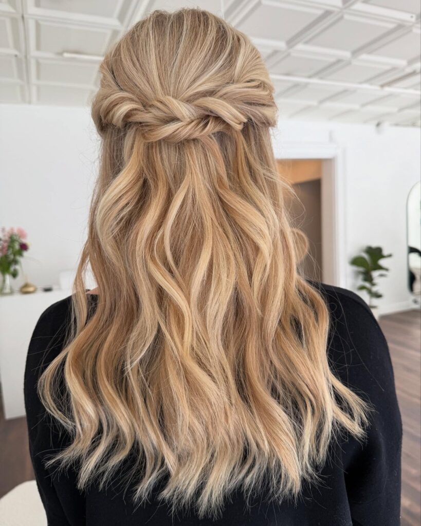A woman with long, wavy, honey blonde hair in a half-updo featuring soft, thick twists at the back.