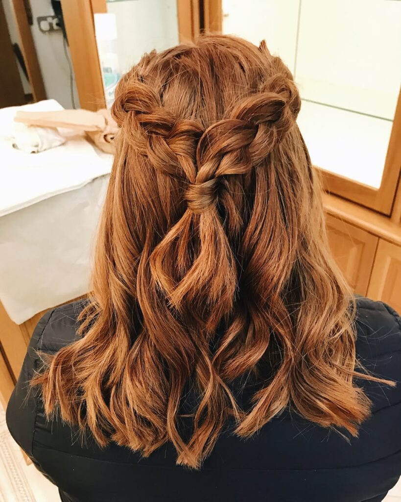 A woman with medium-length auburn hair in a half-updo featuring two thick braids joined at the back with natural waves.
