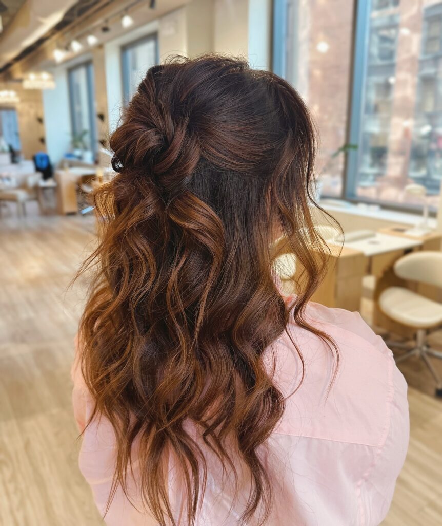 A woman with wavy mahogany brown hair in a relaxed knotted half-updo with soft face-framing pieces.