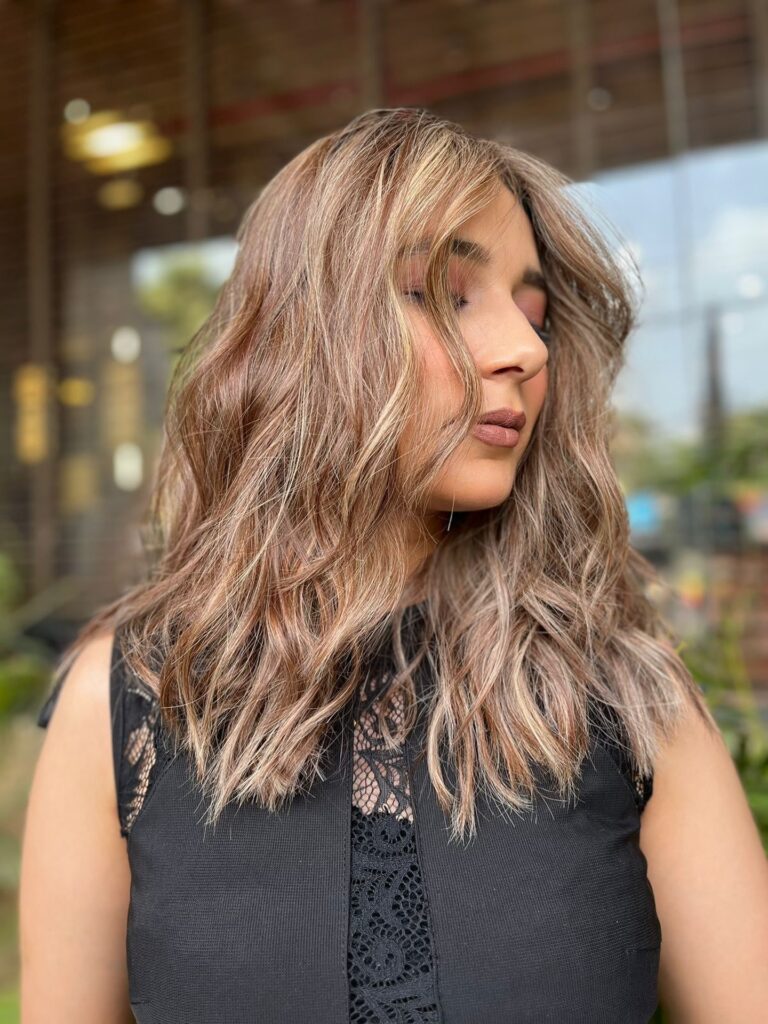 A woman with medium-length wavy hair featuring a blend of honey and caramel blonde highlights.