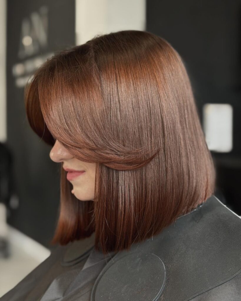 A side profile of a woman with a sleek, blunt, medium-length bob in a glossy copper brown color with a side-swept fringe.