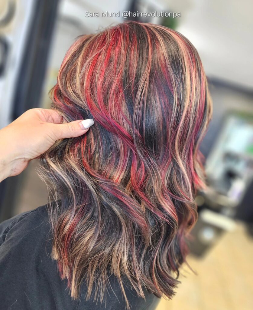 A back view of a woman with medium-length wavy hair featuring a bold mix of red and blonde highlights over dark hair.