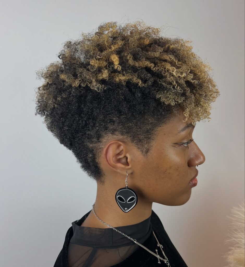 A woman with a short tapered haircut featuring dark natural curls on the bottom and light blonde highlighted curls on top.