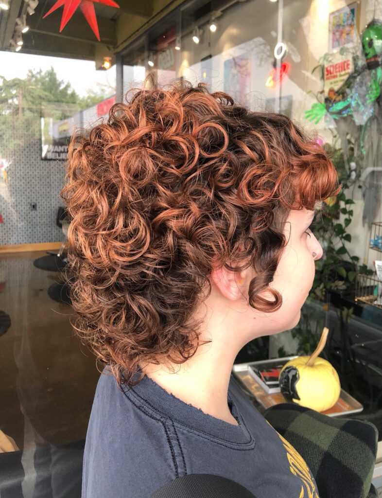 A woman seen from the side with a voluminous curly wolf cut in a vibrant auburn shade.