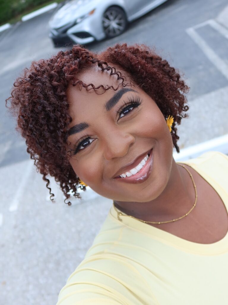 A smiling woman wearing a yellow shirt shows off her short mahogany colored hair with tight natural ringlets.