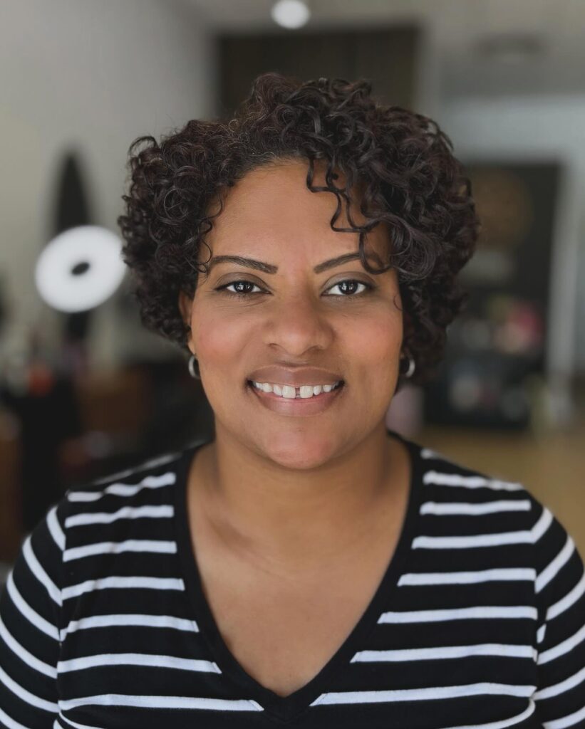 A smiling woman with a short and uniform haircut featuring very defined dark brown natural curls.