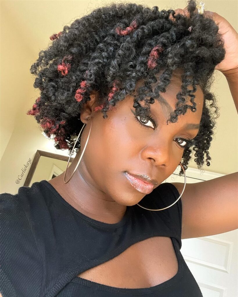 A woman with voluminous chin-length black corkscrew curls featuring several bright red highlighted coils throughout.