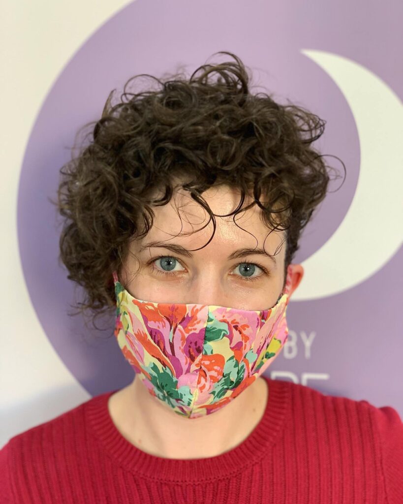 A woman with blue eyes and a face mask wearing her dark curly hair in a short, asymmetrical pixie cut.