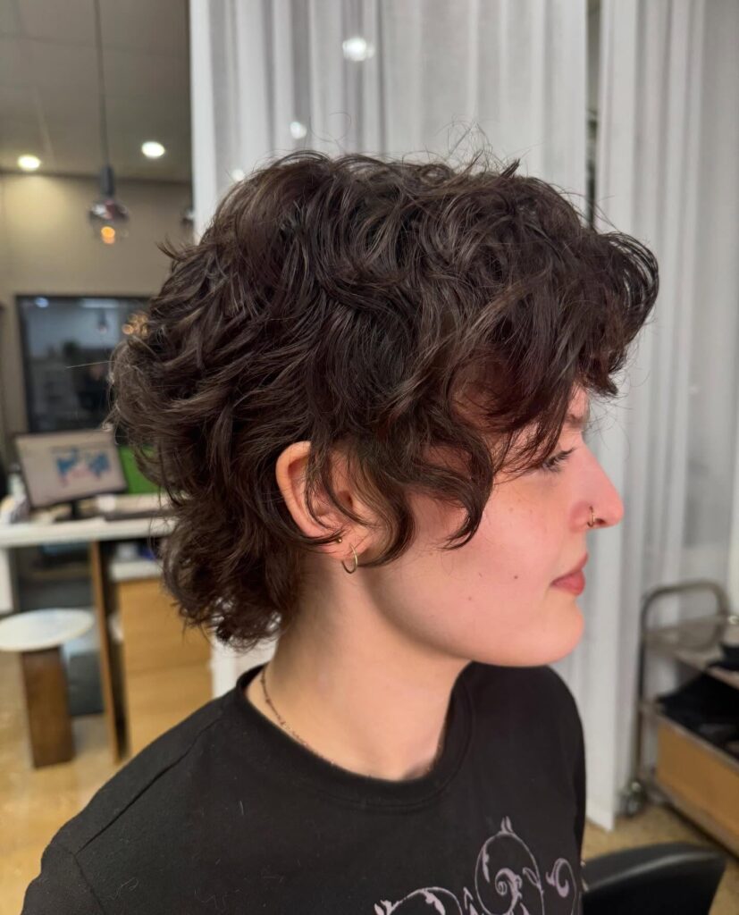 A person seen in profile with a short, shaggy haircut featuring wavy dark brown hair and a soft fringe.