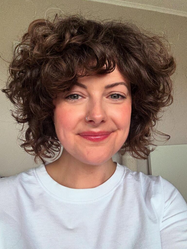 A smiling woman with a voluminous neck-length curly bob in dark brown with curly bangs.