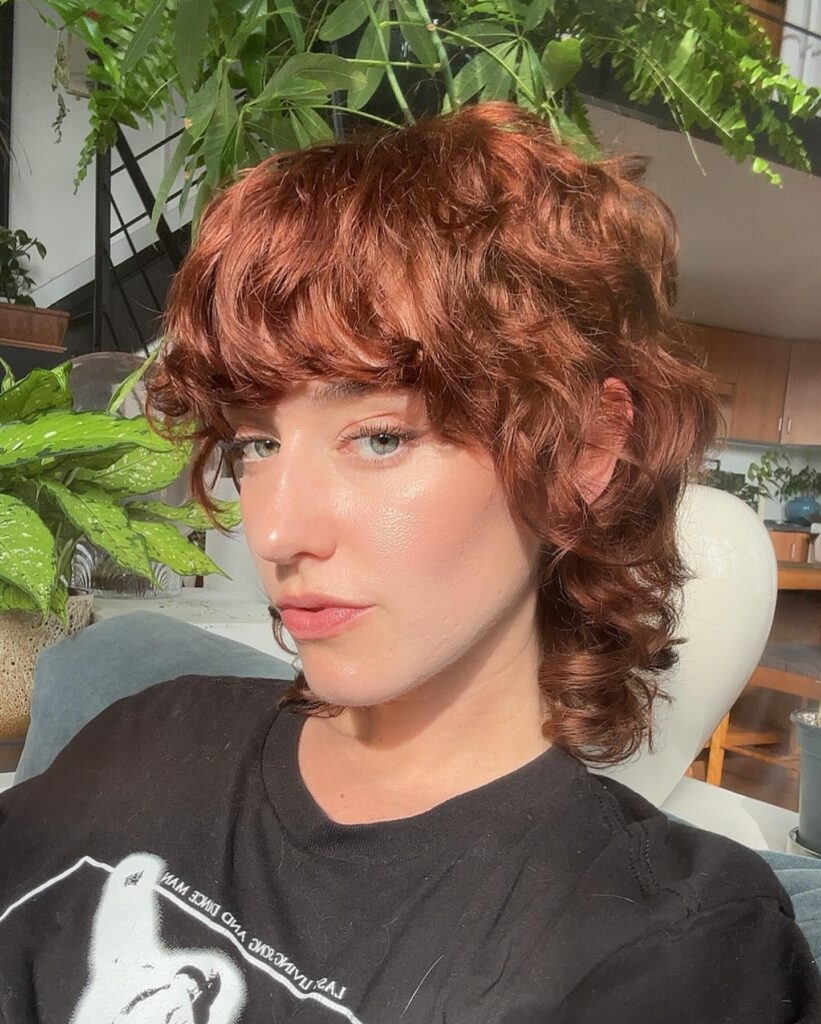 A woman with green eyes and a vibrant copper curly mullet haircut with thick bangs.