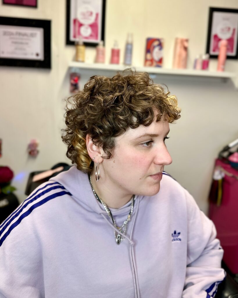 A person with a short curly pixie cut featuring light brown hair and golden blonde peekaboo highlights.