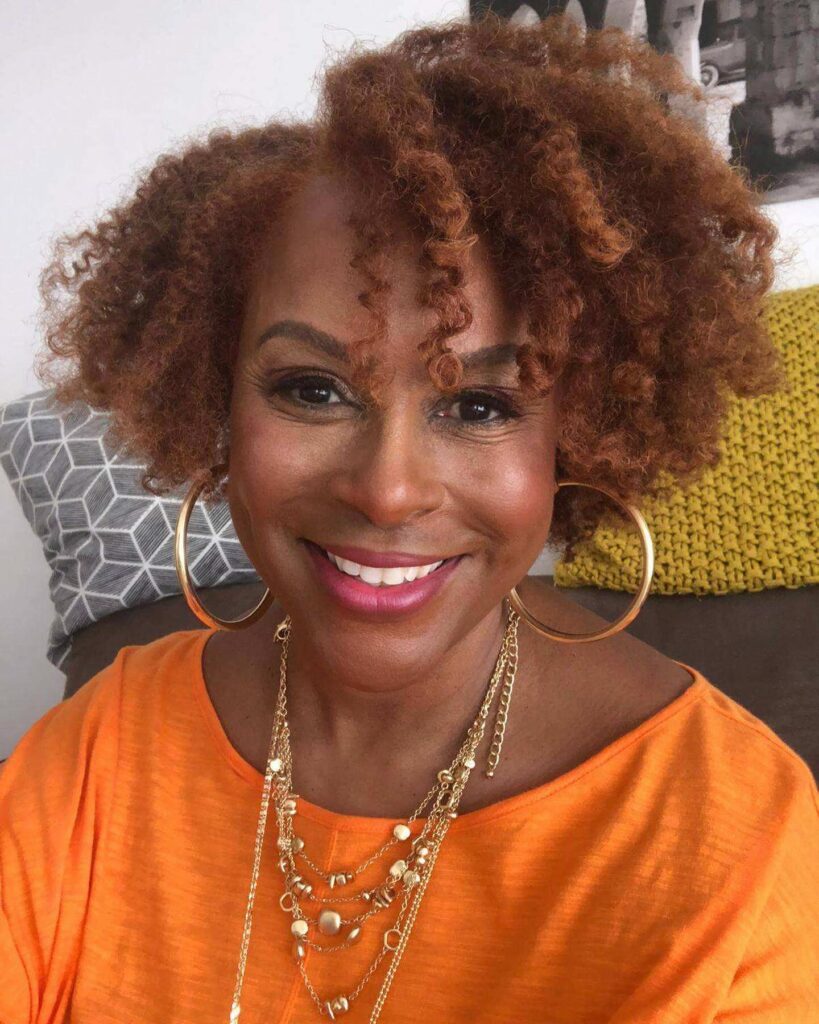 A smiling woman with a voluminous tapered afro featuring tight natural curls in a warm copper-brown color.