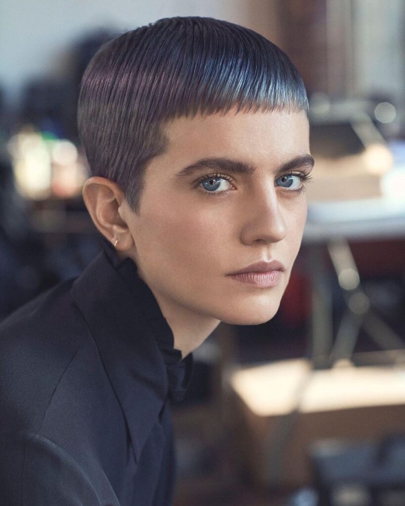 A person with blue eyes sporting an ultra-short pixie cut and micro fringe dyed with iridescent blue and violet tones.