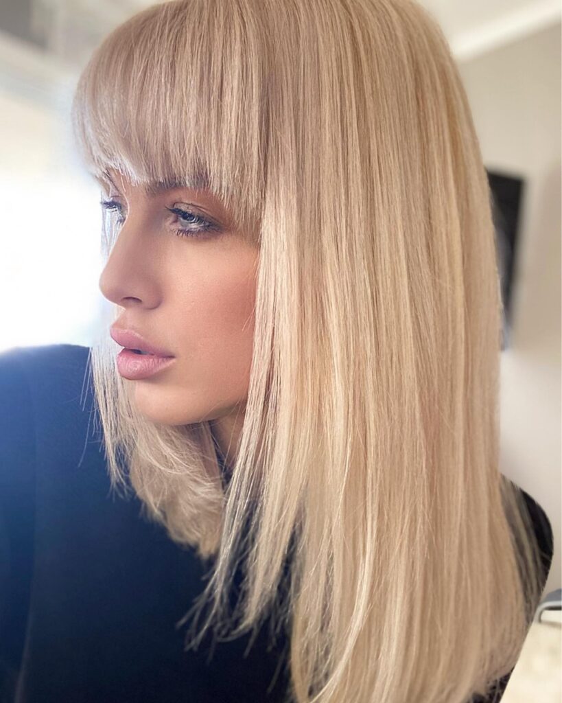 A woman with long, straight, champagne-blonde hair and thick blunt bangs that reach her eyebrows.